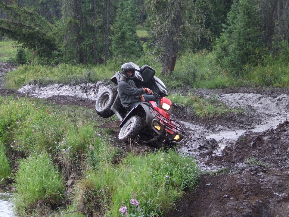 Red ATV with a rider in a helmet navigating a steep, muddy, off-road trail with trees in the background