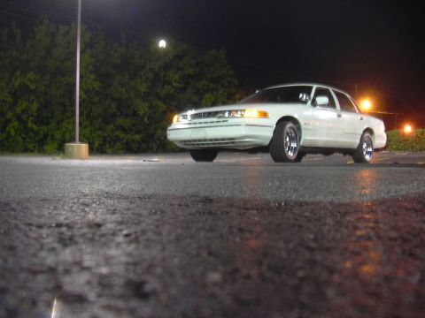 1997 Ford Crown Victoria - A white 1997 Ford Crown Victoria sedan is parked on a wet asphalt surface at night, illuminated by streetlights.