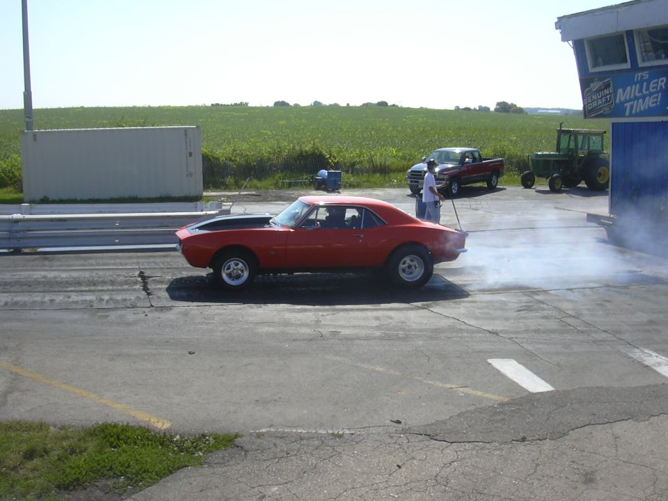 Red 1967 Chevrolet Camaro doing a burnout on a drag strip with a pickup truck and tractor in the background