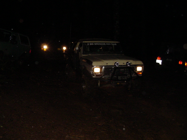 Tan Ford truck with round driving lights and club decal at night, headlights on among other rigs