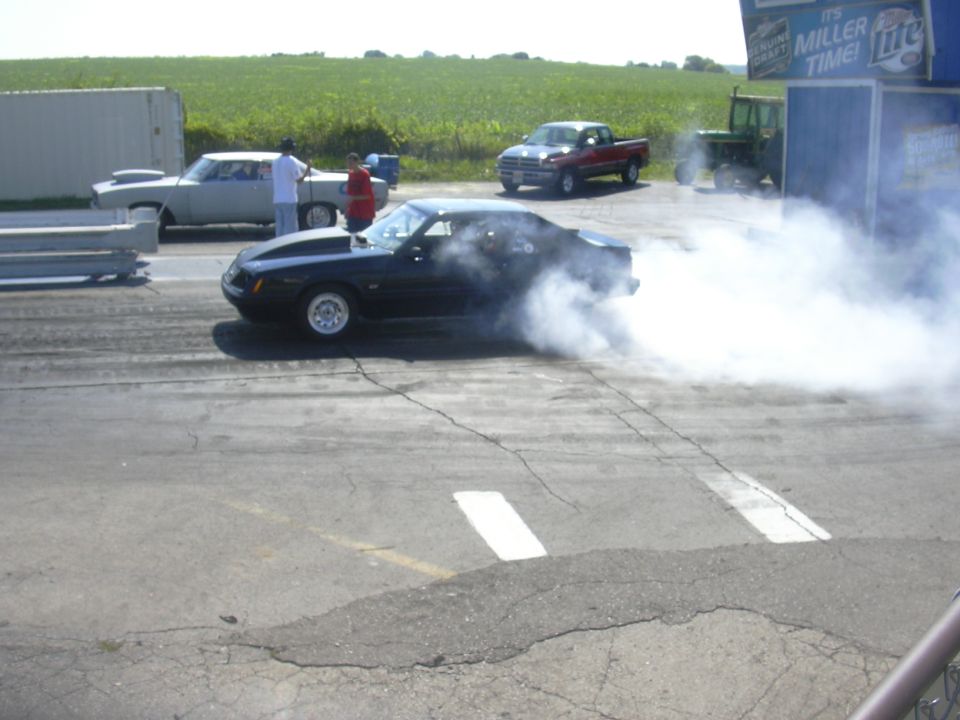 Black Ford Mustang doing a burnout on a drag strip, with a light-colored sedan and a red pickup truck in the background