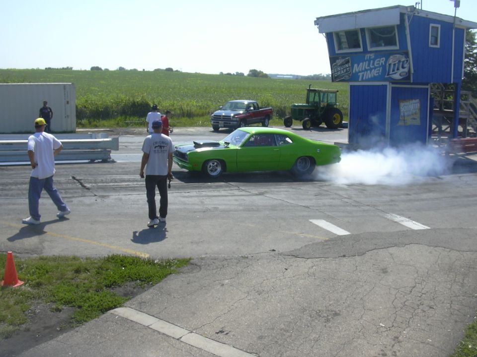 Green muscle car doing a burnout on a drag strip, with a tractor and pickup truck in the background