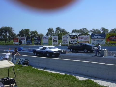 1997 Ford Crown Victoria - Two cars prepare for a drag race at a track with numerous sponsor advertisements visible.