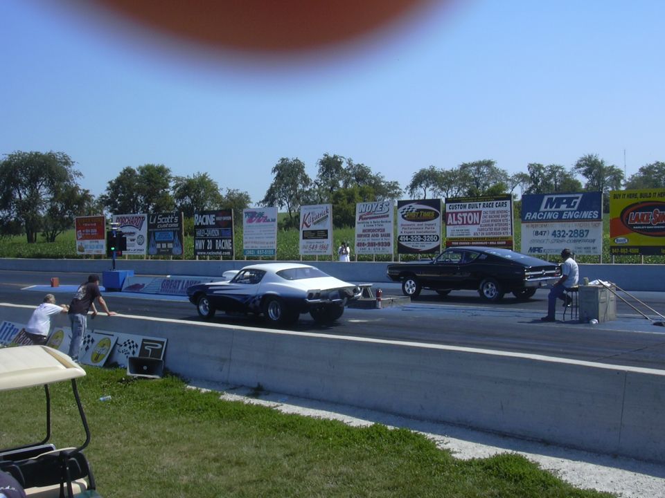 two muscle cars lined up on a drag strip, one black, one white and black, with spectators and sponsor signs in background