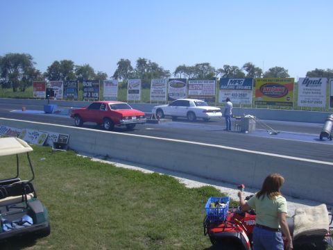 1997 Ford Crown Victoria - A white sedan is positioned on the drag strip, with a red car to its left and a person standing nearby.