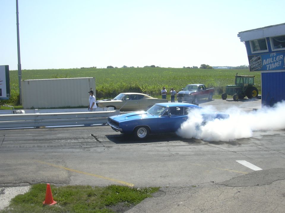 Blue muscle car doing a burnout on a drag strip, with a tan car and a red truck behind it