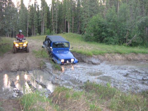 1995 Ford Bronco - A blue Jeep Wrangler is stuck in a muddy rut while an ATV waits on a dirt track nearby.