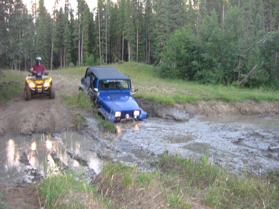 Blue Jeep Wrangler partially submerged in mud with an ATV on a dirt path behind it