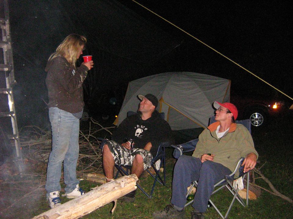 People gathered around a campfire at night with a tent and a vehicle in the background