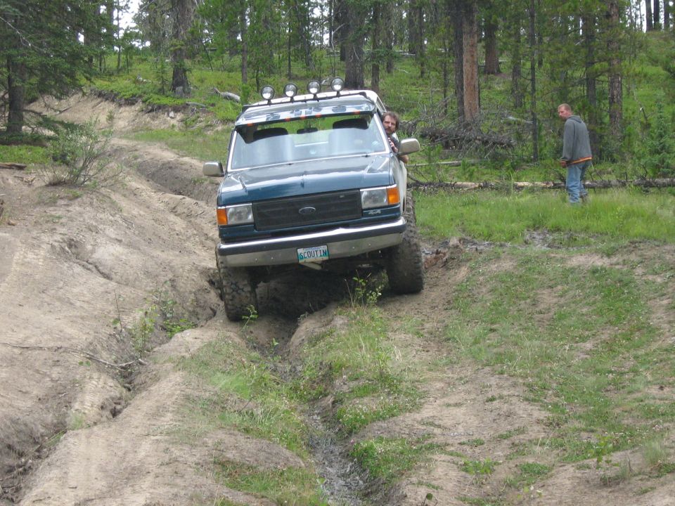Dark green Ford Bronco on a dirt trail, front-left wheel in a ditch, roof lights, driver visible