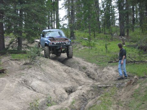 1995 Ford Bronco - A man observes a modified blue truck navigating a challenging dirt path through a forest.