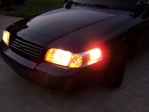 2003 Ford Crown Victoria - The front corner of a dark 2003 Ford Crown Victoria is visible with its headlights and turn signals on.