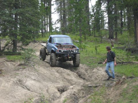 1995 Ford Bronco - A modified blue pickup truck navigates a challenging dirt path in a wooded area.