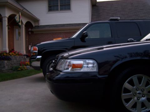 2003 Ford Crown Victoria - Two black vehicles, a sedan and an SUV, are parked in a driveway in front of a house.