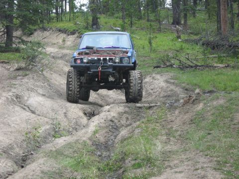 1995 Ford Bronco - An off-road vehicle navigates a challenging dirt trail in a wooded area, its hood open.