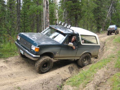 1995 Ford Bronco - A Ford Bronco navigates a rough dirt path in a wooded area, with another off-road vehicle following.