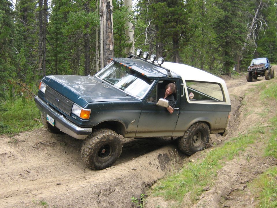 Green Bronco with white top driving on a dirt trail, another vehicle behind it