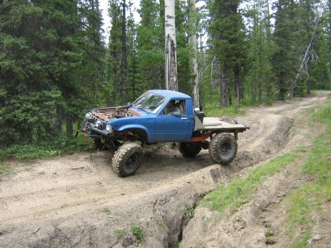 1995 Ford Bronco - Modified blue flatbed truck tackles a challenging off-road trail with deep ruts and steep inclines.