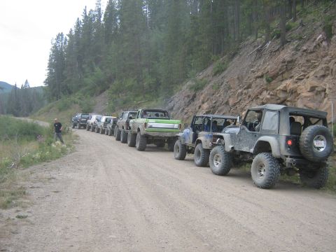 1995 Ford Bronco - Multiple off-road vehicles are lined up on a dirt road, heading into a wooded, mountainous region.