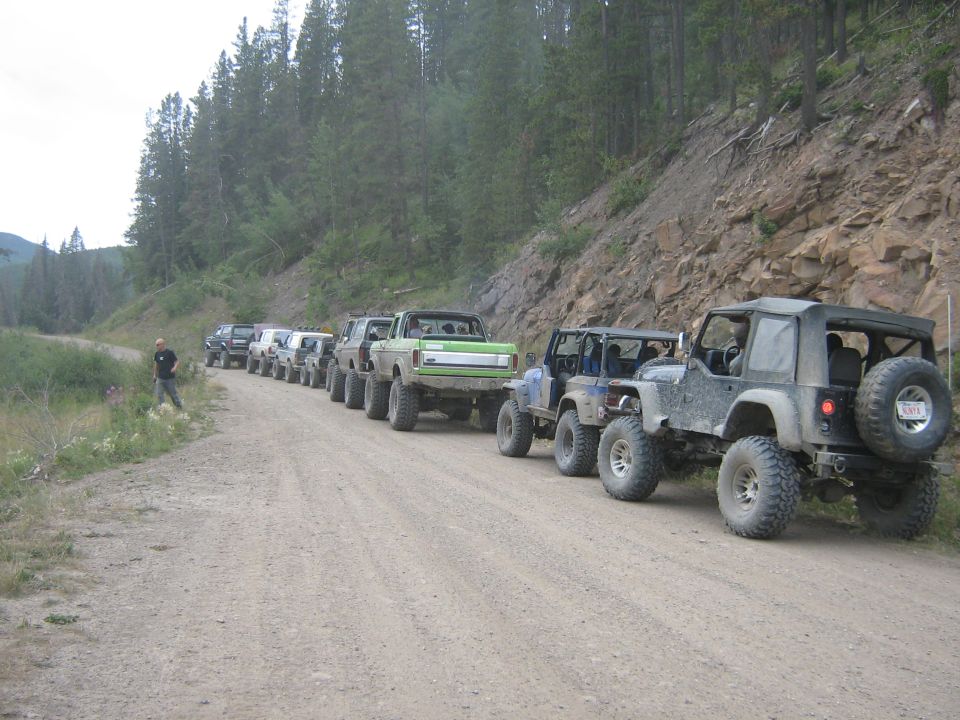 Line of off-road vehicles including Jeeps and a green truck on a dirt road in a mountainous area