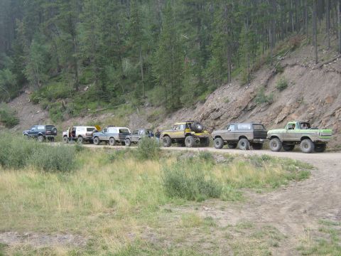 1995 Ford Bronco - Several off-road vehicles are lined up on a dirt road in a wooded area.
