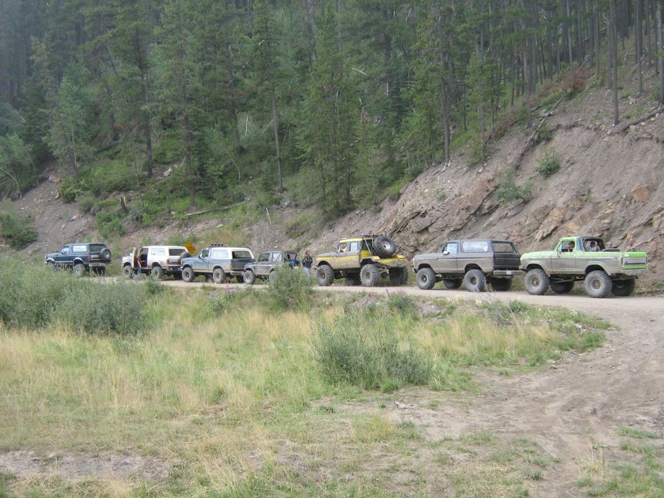 Line of off-road SUVs and trucks parked on a dirt road next to a wooded hillside