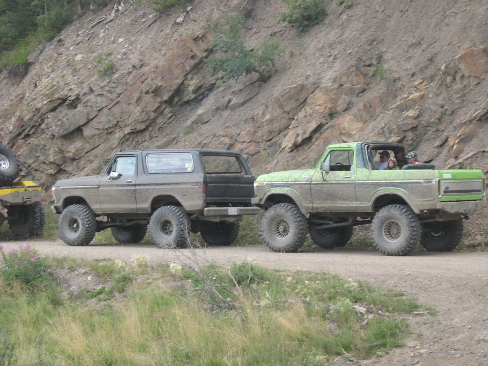 Two lifted Ford Broncos, one grey and one green, on a dirt road with a rocky hillside