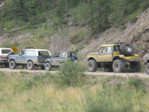 1995 Ford Bronco - Several modified off-road vehicles, including a 1995 Ford Bronco, travel on a dirt road through a wooded, mountainous area.