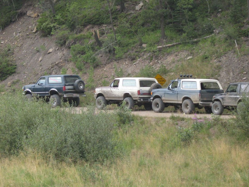 Several Ford Broncos driving on a dirt road in a wooded area. One Bronco is dark blue, others are white and gray