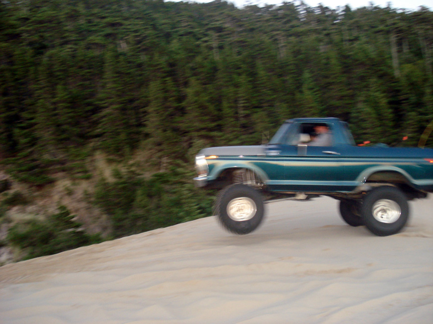 1979 Ford Bronco lifted and driving on a sand dune with trees in the background