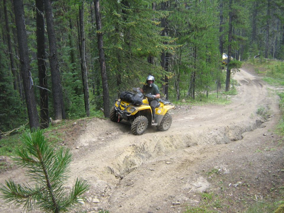 Yellow ATV with rider on a muddy, rutted trail in a forest. Another ATV is visible further up the trail