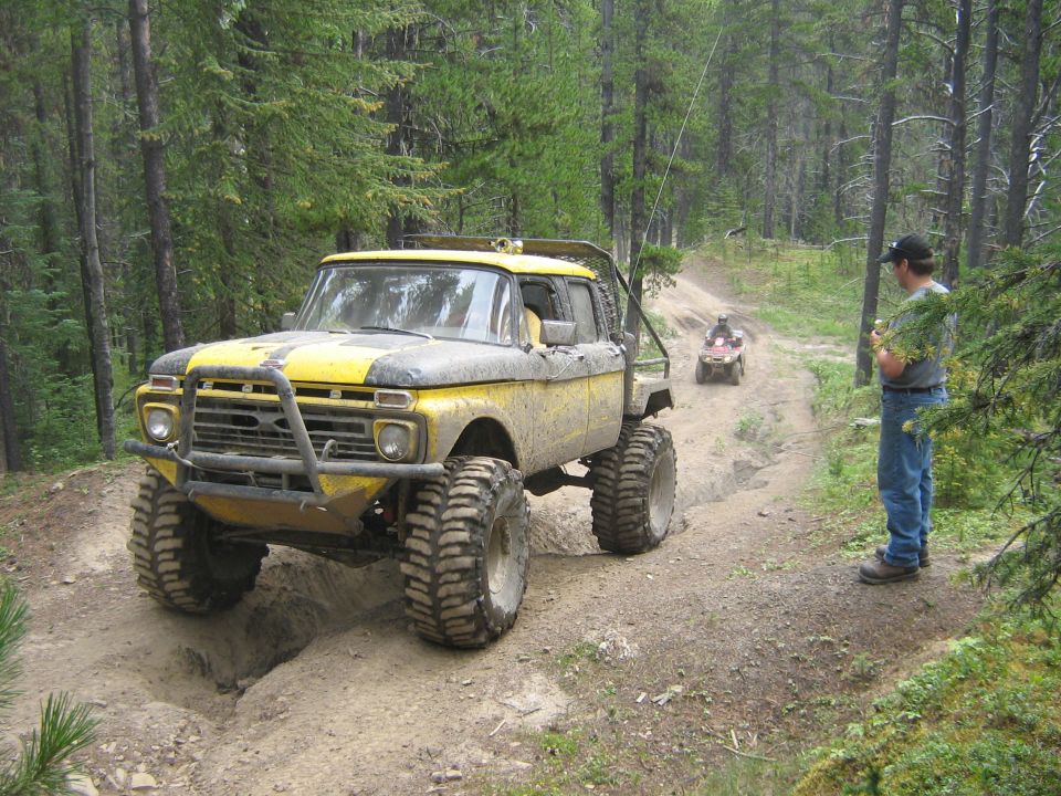 Yellow and grey lifted truck with large tires on a dirt trail, a person stands nearby