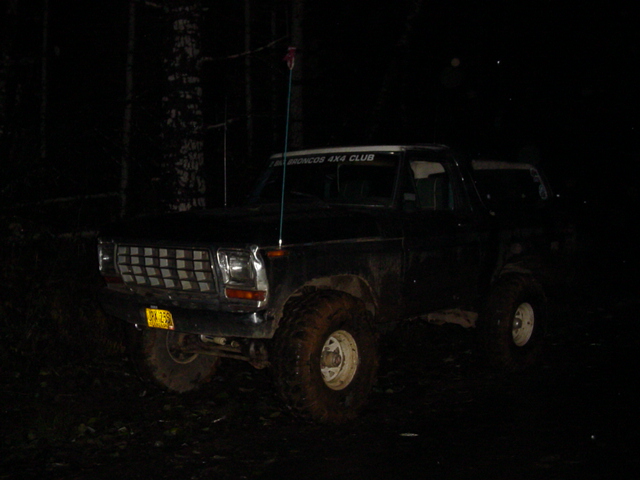 Dark older Ford Bronco with whip antenna and club decal parked among trees at night
