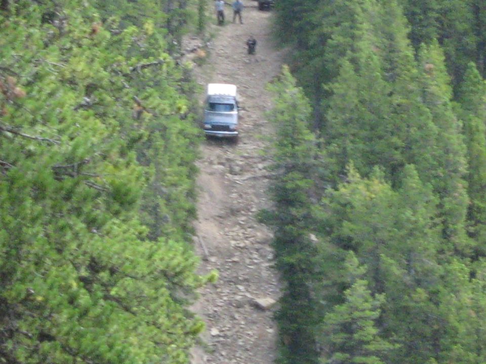 Van driving up a steep, rocky dirt road through a forest. People are visible on the road ahead