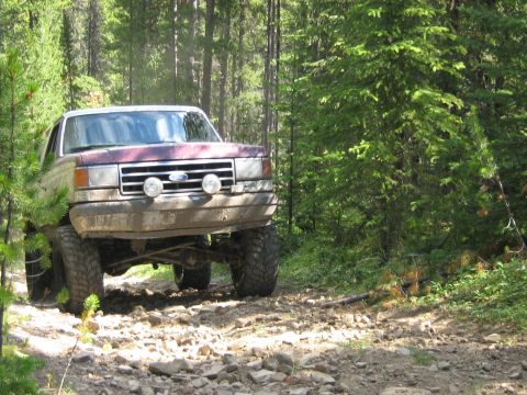 1995 Ford Bronco - Maroon and white Ford F-Series truck navigates a rocky path through a dense forest.