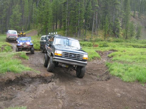 1995 Ford Bronco - Several off-road vehicles, including a Ford Bronco, traverse a challenging muddy track in a wooded area.