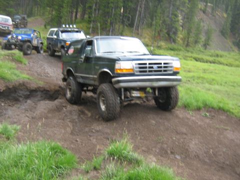 1995 Ford Bronco - Several off-road vehicles navigate a challenging dirt path in a wooded area.