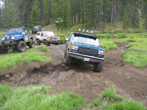 1995 Ford Bronco - A teal 1995 Ford Bronco tackles a challenging muddy obstacle during an off-road rally, with other vehicles following behind.
