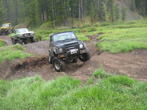 1995 Ford Bronco - Suzuki Samurai tackles a challenging off-road course during a rally event, with other trucks and SUVs following behind.