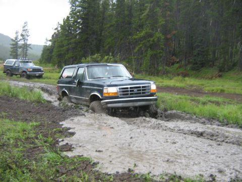 1995 Ford Bronco - Two Ford Broncos navigate a muddy trail with trees in the background.