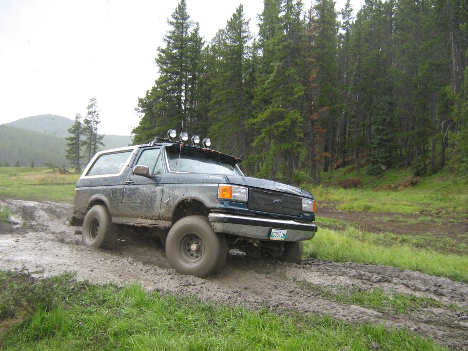 Dark green 1992 Ford Bronco driving through mud, front right tire visible, roof lights on