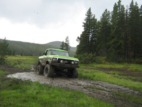 1995 Ford Bronco - Off-road truck navigates a muddy trail surrounded by trees and grassy fields.
