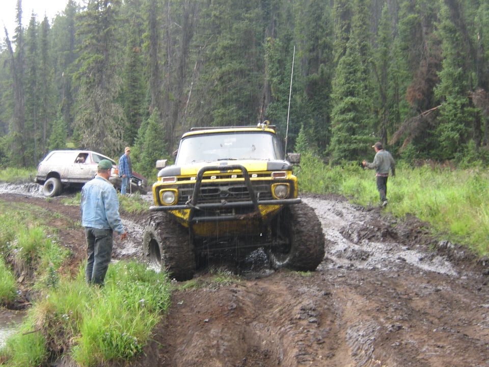 Yellow off-road truck with large tires stuck in mud, white SUV behind it, forest background