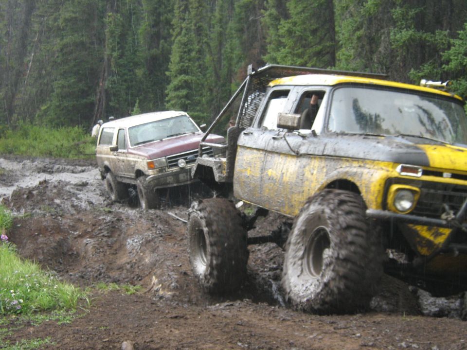 Two mud-covered 4x4 trucks stuck in deep mud on forest trail