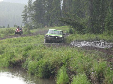 1995 Ford Bronco - Two off-road vehicles navigate a wet and muddy trail through a wooded area.