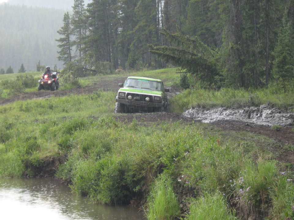 Green lifted 4x4 truck in mud bog beside ATV on grassy forest trail