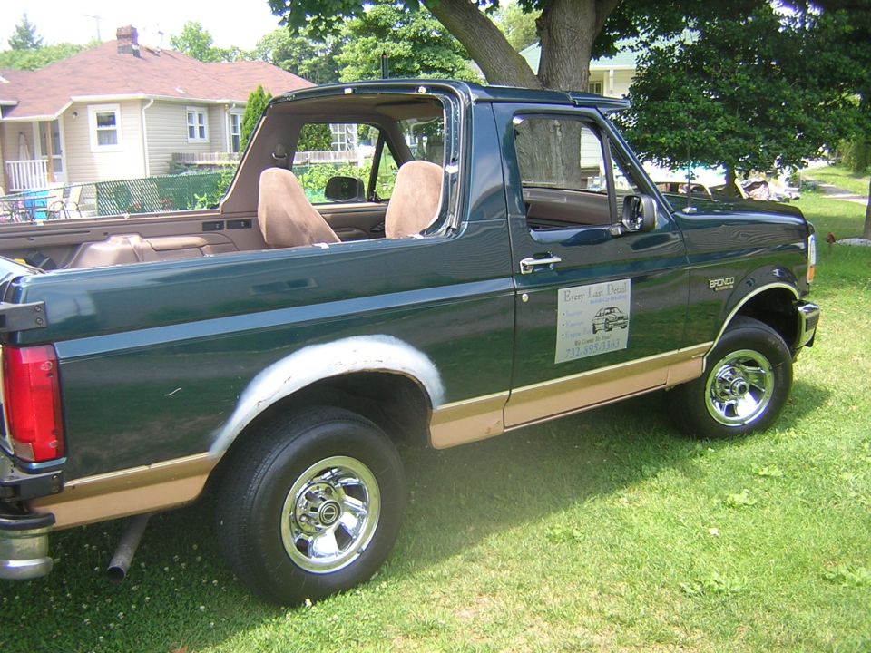 Dark green Ford Bronco with top removed, tan two-tone trim, primer patches on rear fender, parked on grass