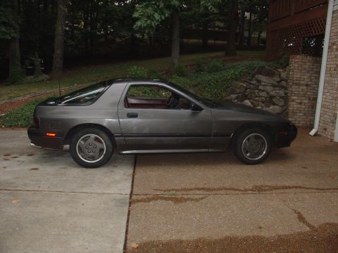 1986 Mazda RX-7 - A gray 1986 Mazda RX-7 is parked on a concrete driveway next to a brick house.