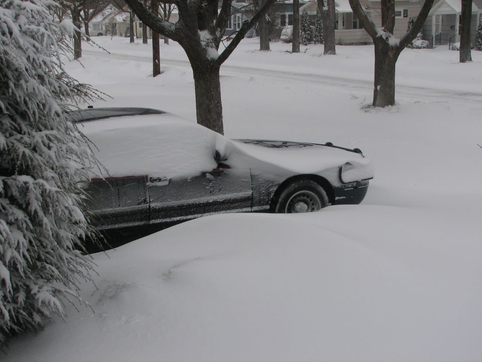 Black sedan buried in deep snow in a driveway, only side and front wheel visible