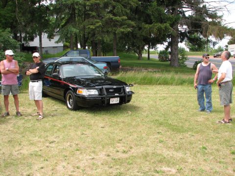 2000 Ford Crown Victoria - A group of men stand around a black Ford Crown Victoria parked on a grassy area, with trees and houses in the background.